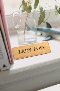 bronze plaque with words lady boss, on a window sill beside a pink book and a glas vase with a green leaf in it