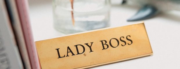 bronze plaque with words lady boss, on a window sill beside a pink book and a glas vase with a green leaf in it