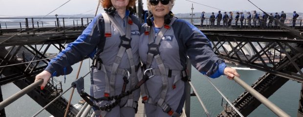Two women in sunglasses and grey and blue overalls on top of Sydney harbour bride holding wire railing with metal tower and australian flag in background. Green water below.