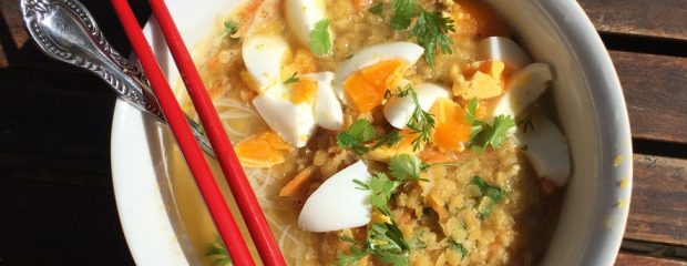white bowl containing red lentil soup, rice noodles and quartered boiled egg garnished with coriander a spoon sits in the bowl and a pair of red chopsticks across the top