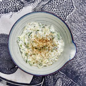 view of blue rimmed bowl containing yoghurt, mint, cucumber raita topped with toasted cumin seed on black and white tea towel