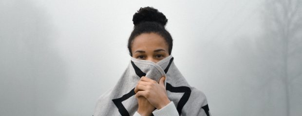 woman of colour with dark hair in a bun at top of head holds gray and black geometric striped scarf of pbdy and lower face background white mist it looks cold