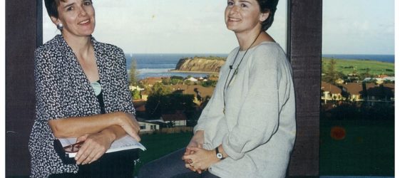 two women sit on a wall smiling with sea in the background