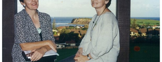 two women sit on a wall smiling with sea in the background