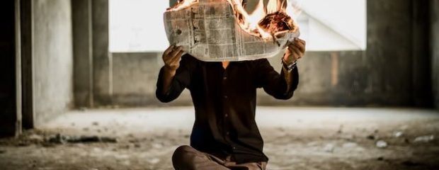 man in dark suit sits with leg crossed over knee, newpaper on fire blocks his face