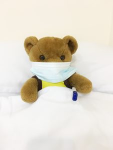 Teddy in bed tucked up with white sheets wearing a surgical mask with blue and white digital thermometer under arm