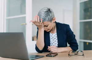 Middle aged woman with short grey hair sits at desk and open laptop with head in her hand looking exhausted is she anaemic?